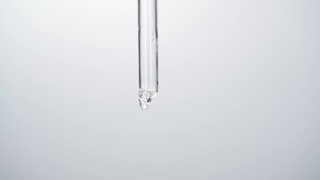 Macro close up of glass dropper pipette dripping clear and pink cosmetic liquid serum drop on white background, science laboratory and skincare concept