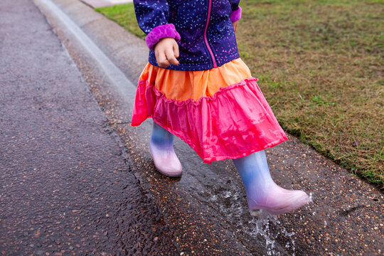 child in raincoat and boots splashing through roadside gutter full of water on rainy day