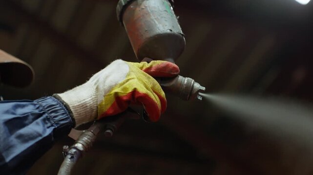 Gloved worker spraying paint onto metal parts with handheld spray gun inside industrial workshop during surface coating and finishing process under warm directional light