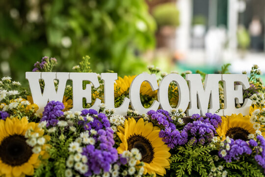 Bright white wooden welcome sign lettering placed carefully among a vibrant arrangement of yellow sunflowers and purple statice flowers in a sunny garden setting captured with a soft focus effect