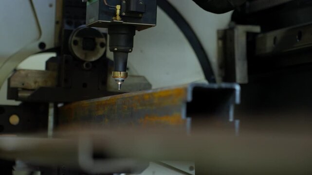 Moving laser cutting head throws bright sparks across steel channel edge inside industrial fabrication machine, showing precise thermal cutting motion in workshop