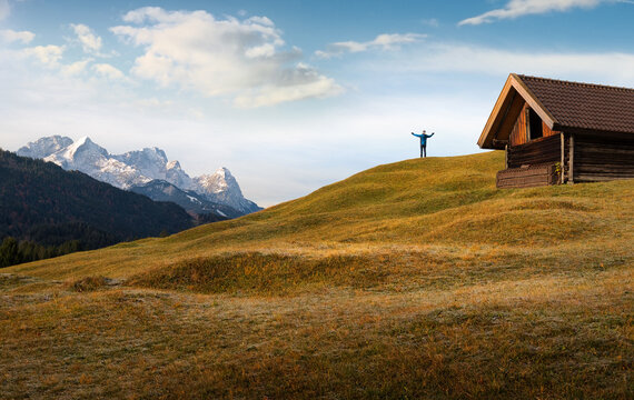 Man standing on the edge of a mountain in front of a mountain cabin and greeting the vastness of the alpine landscape
