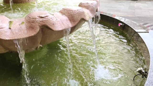 Serene Stone Shell Fountain Water Flow. Gentle streams of crystal clear water pour from a stylized stone shell basin into a shimmering green urban pond pool. Video transition, 4k, 60 fps