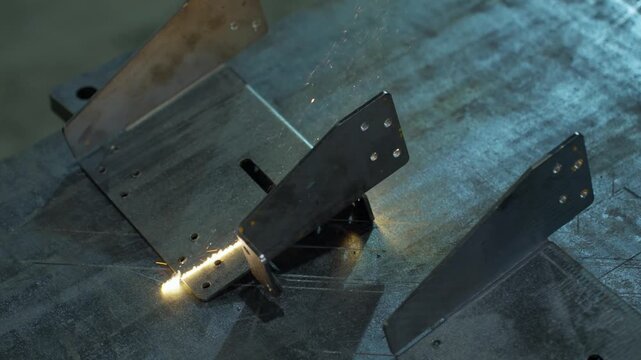 Close-up laser metal processing on fixture with bright molten seam and sparks, showing reflective sheet and steel brackets on scratched worktable in workshop, with laser, metal, cutting, spark detail