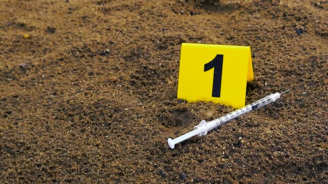 Yellow forensic evidence marker with the number one next to a used syringe on the wet sand during a rainy day, highlighting a potential crime scene investigation related to drug abuse