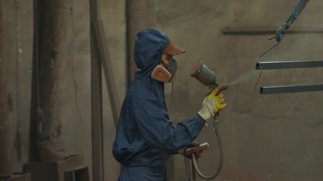 Industrial worker spray painting suspended metal bars inside workshop while wearing protective respirator, hooded coverall, cap, and gloves during coating process