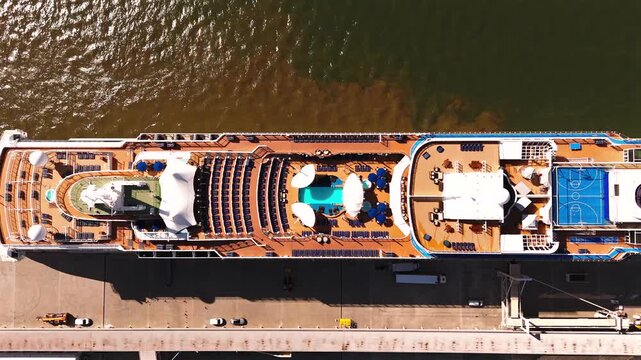 A drone looks straight down on a white cruise ship with pools and sun decks beside a calm harbor