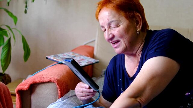 woman aged 70- 75 sits near houseplants and speaks on speakerphone while the device is plugged into a wall outlet. The scene can illustrate everyday communication, senior technology use.