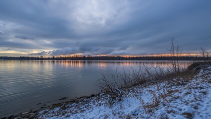 Fototapeta premium Winter Sunset Over Calm Lake with Snow-Covered Shoreline and Dramatic Clouds