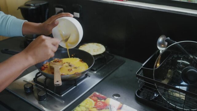 Man cooking, pouring eggs into a pan to make an omelet with onion and tomato, and browning a Colombian arepa