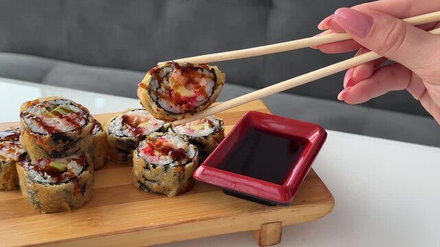 Golden fried sushi rolls are carefully dipped into rich soy sauce using wooden chopsticks above a red rectangular dipping dish