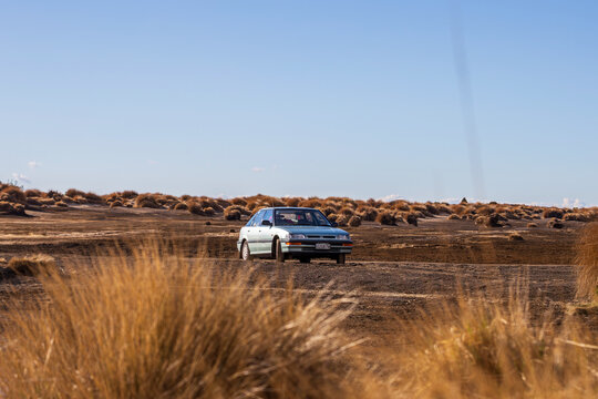 A light blue sedan pauses on the dry, rugged expanse of Desert Road, New Zealand, surrounded by sparse vegetation and a vast open sky