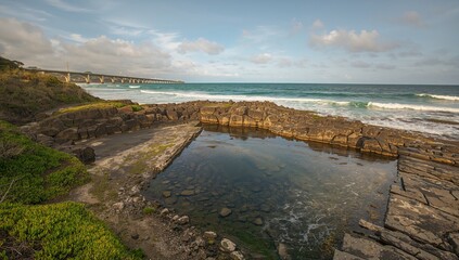 Obraz premium Rocky Coastal Pool with Ocean Waves and Bridge Under Cloudy Sky