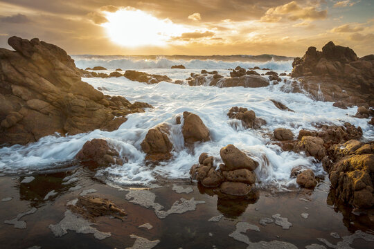 Low angled view of waves washing over rocks along a rugged coastline under a colourful sunset sky
