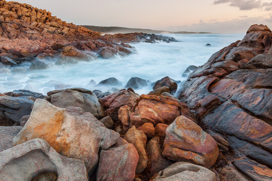 Long exposure of waves washing over colourful granite boulders along a rocky coastline at twilight