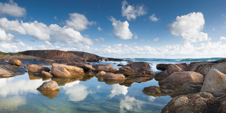 Low angled panorama view of clouds reflected in a coastal rockpool surrounded by granite boulder