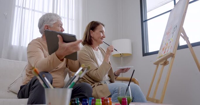 Senior woman paints canvas and checks tablet reference held by husband, discussing ideas and smiling together during creative art therapy, cognitive engagement, companionship, and retirement wellness.