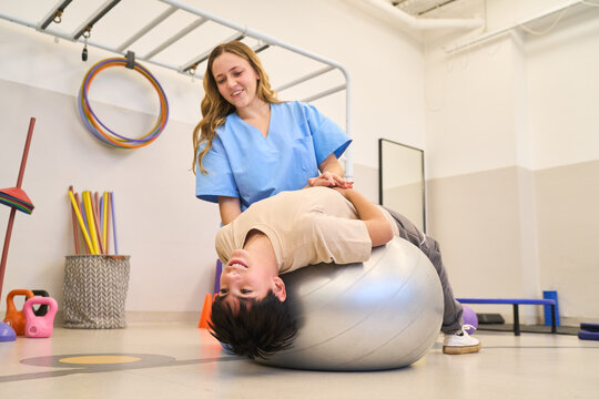 Pediatric physiotherapy session, child rehabilitation exercises with therapist, balance training on exercise ball, motor skills development in pediatric rehab center, happy child