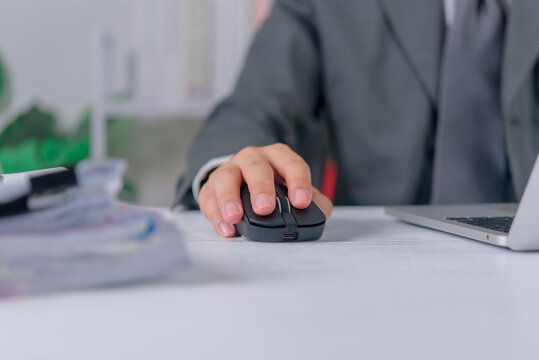 Hand of a businessman in a suit uses a black wireless computer mouse at an office desk. The corporate professional works on a laptop next to a stack of business documents in a bright workplace.