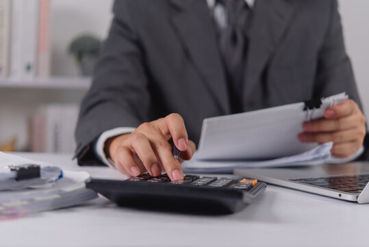 Close up of an accountant in a suit using a black calculator while holding a paper document. The corporate professional calculates business finances and taxes at a busy office desk with a laptop.