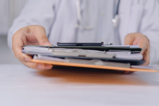 Hands of a medical professional in a white coat holding a large stack of patient record clipboards. The doctor offers or passes clinical files and health paperwork across a bright clinic desk.