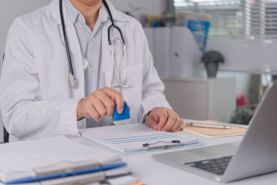 Male doctor in white lab coat uses a blue rubber stamp to authorize clinical documents on a clipboard. The healthcare professional approves patient paperwork at a modern clinic desk with a laptop.