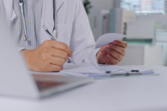 Hands of a physician in a white lab coat writing on clinical documents with a pen. The medical professional signs a prescription or patient report on a clipboard at a bright office workspace.