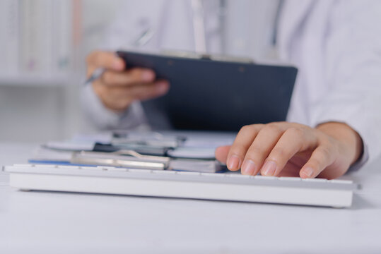 Hands of a doctor in a white lab coat typing on a computer keyboard at a clinic desk. The medical professional updates electronic health records and clinical data with a clipboard in background.