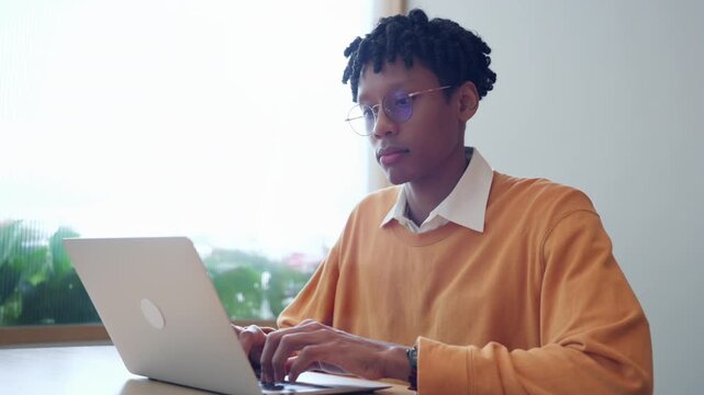 Focused young black man typing on laptop while working remotely in bright modern office.