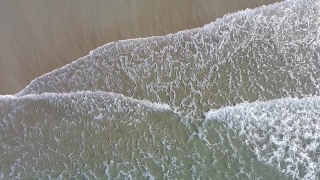 15 second aerial footage from a drone looking down on waves gently washing up on a sandy beach in the summer. 