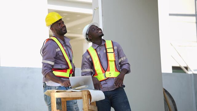 Two black construction worker wearing safety vest and hardhat look at laptop on wooden table together to discuss residential real estate building project on site