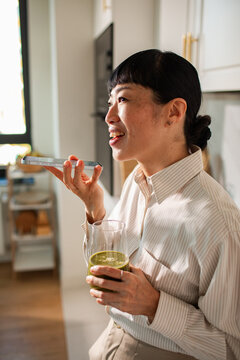 Smiling woman using voice assistant on smartphone at home kitchen with green smoothie