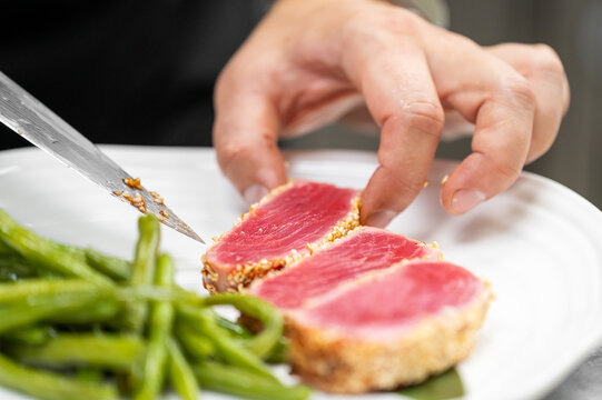 A chef's hand carefully places sliced seared ahi tuna with sesame seed crust on a white plate, served with green beans.