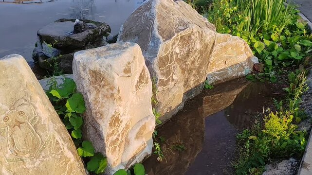 Ancient stone carvings with frog design by a tranquil water body surrounded by vegetation