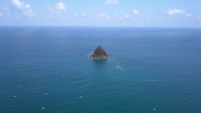 Aerial view of rocky islet surrounded by fishing boats in blue ocean waters.