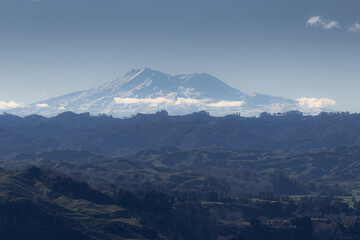 Fototapeta premium A majestic view of Mount Ruapehu, New Zealand, with snow-capped peaks under a clear blue sky. The foreground shows rolling hills covered in lush green vegetation.