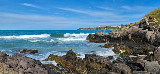 Fototapeta premium Waves crashing on a rocky coastline with a distant cliffside town under a clear blue sky.