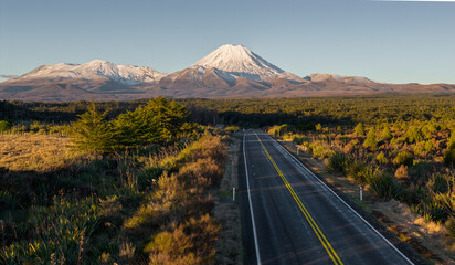 Obraz premium A lonely road winds through the Tongariro National Park in New Zealand, with Mount Ngauruhoe looming in the distance. A caution sign warns of kiwi crossings at night.
