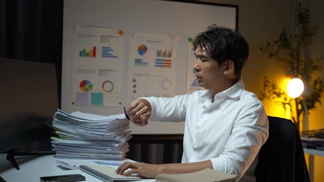 Stressed businessman working late at night feeling neck pain and office syndrome from overworking with a pile of paperwork and documents on his desk in a dark office