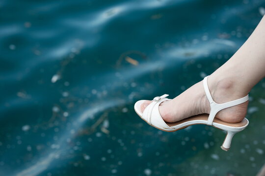 Close-up of a woman's foot wearing a white low-heeled sandal dangling over blue sea water.