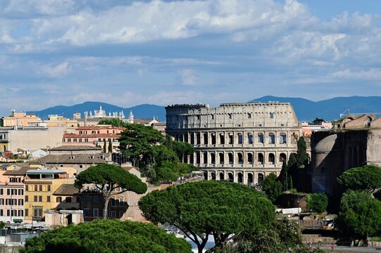 scenic aerial view of the Colosseum arena building in Rome, Italy. old brick and stone buildings around. lush green pine trees. blue sky and white clouds. mountains in background. architecture, travel