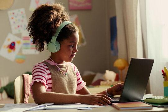 Black girl wearing headphones using laptop for online learning and homework at desk, showing digital education and child focus. Useful for e learning, homeschooling, edtech campaigns