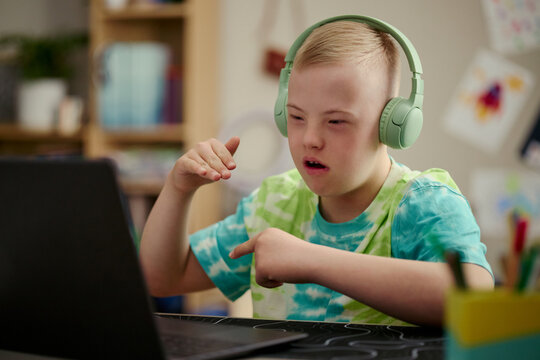 Caucasian child with Down Syndrome wearing headphones using laptop and gesturing during online lesson, showing inclusive learning and communication. Ideal for education support campaigns