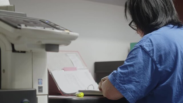 Side rear view of a medical administrative worker managing records at a desktop workstation. Suitable for healthcare administration, clinical documentation, and office workflow.