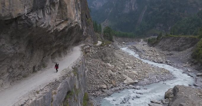 Annapurna Circuit, Manang region, Nepal, aerial pull-out shot revealing the sheer scale of a mountain cliff trail with trekkers hiking high above the rushing Marsyangdi River.