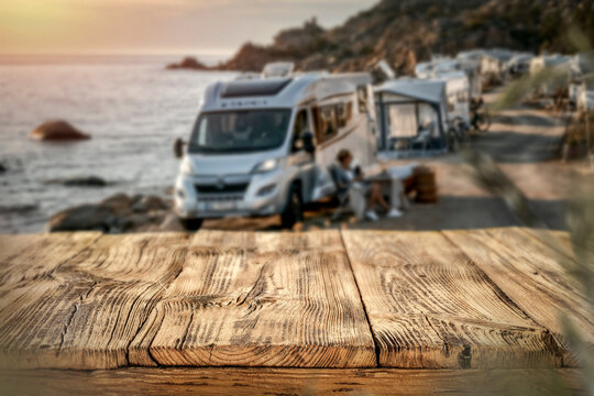 Copy space on empty wooden table with the background of camping scene with camper van by rocky seaside &ndash; summer travel and outdoor adventure concept.