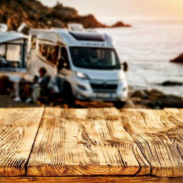 Copy space on empty wooden table with the background of camping scene with camper van by rocky seaside &ndash; summer travel and outdoor adventure concept.