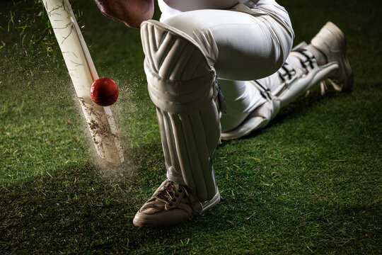 Close-up cricket bat stopping red ball beside batter leg pad on grass field. Concept of precise batting, defensive technique, cricket equipment, sports marketing and commercial campaigns.