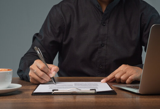 Person signing paperwork on clipboard beside laptop