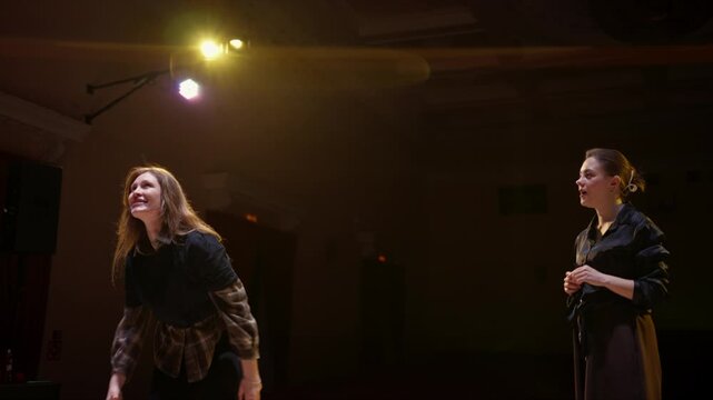 Two women rehearsing a dramatic performance on a dimly lit stage, practicing expressive movements and body language under warm theatrical spotlight lighting